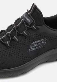 Black athletic shoe with mesh fabric, elastic lacing, and textured sole. Features a prominent Skechers logo on the side.