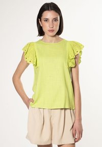 Limegrøn kortærmet top med øjelet flæseærmer, struktureret stof og en afslappet pasform, parret med lyse beige shorts.