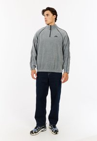 Grauer Fleece-Pullover mit hohem Kragen und Halbreißverschluss, akzentuiert mit schwarzen Nähten. Kombiniert mit dunkelblauen Jeans und gemusterten Schuhen.