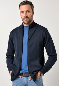 Granatowy kardigan na zamek z prążkowanym kołnierzem, teksturowaną tkaniną, dwoma przednimi kieszeniami, noszony na niebieskiej koszuli, zestawiony z jasnoniebieskimi dżinsami.