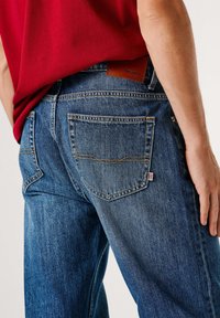 Vaqueros de mezclilla azul con un diseño clásico de cinco bolsillos. La parte trasera presenta costuras decorativas y una etiqueta de cuero marrón con la marca.