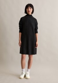 Črna pletena obleka z visokim ovratnikom, dolgimi rokavi in ravnim krojem, v kombinaciji z belimi supergami in rebrastimi manšetami. Gladka tekstura.