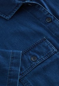 Camisa de mezclilla azul con cuello puntiagudo, dos botones negros y costuras en contraste. La tela tiene una textura suave con un ligero brillo.