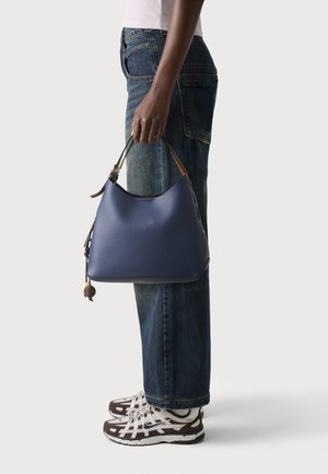 Personne portant un jean bleu et des baskets tient un sac à main en cuir bleu avec une poignée marron, debout devant un fond uni.