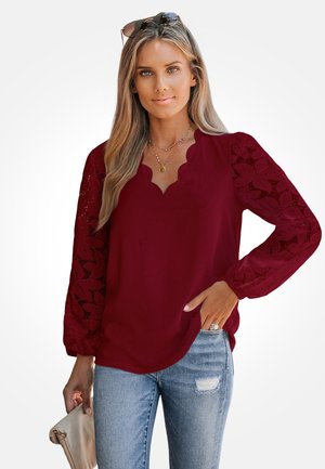 Blusa color burdeos con escote en forma de ondas y mangas largas de encaje con un patrón floral. Combinada con jeans desgastados de lavado claro.