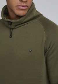 Felpa verde oliva con colletto alto, chiusura con cordino e patch logo nero. Texture liscia e design aderente con maniche raglan.