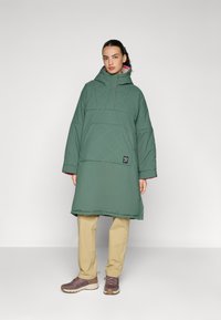 Abrigo acolchado verde y oversize con cuello alto, forro interior rosa, gran bolsillo frontal, llevado sobre pantalones color beige y zapatos marrones.