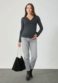 Έγκυος γυναίκα φορώντας μαύρη μπλούζα μακρύ μανίκι και γκρι τζιν, κρατώντας μια μαύρη τσάντα με χρυσή αλυσίδα, στέκεται σε γκρι δάπεδο.