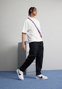 Fehér, oversized póló, fekete cargo nadrág és fehér Nike sportcipő fekete swoosh-sal. Kék és piros heveder a vállon.