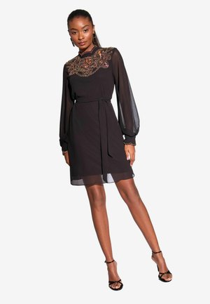 Rochie neagră cu mâneci lungi din material transparent, guler înalt și broderie colorată complicată pe corset. Include o talie legată și o fustă stratificată.