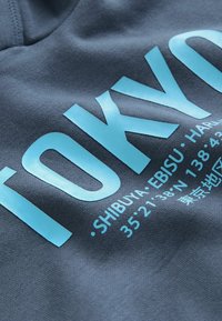 Tissu gris foncé avec un texte imprimé bleu clair portant "TOKYO" et des coordonnées de localisation, affichant une texture lisse et un design graphique.