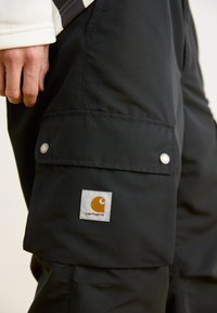 Crne cargo hlače izrađene od izdržljivog materijala, s bočnom džepom sa srebrnim napinjem i bijelom etiketom s narančastim logom.