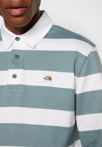 Homme portant un polo de rugby à rayures horizontales bleu-gris et blanches avec un col blanc et le logo The North Face sur la poitrine.