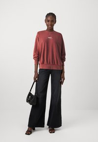 Felpa rossa con collo a giro e orlo elastico, abbinata a jeans neri a gamba larga a vita alta. Il modello tiene in mano una piccola borsa nera.
