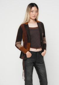 Bruine corduroy blazer met meerkleurige panelen, beige en roestkleurige accenten, gecombineerd met een donkerbruine top en zwarte gemêleerde broek.