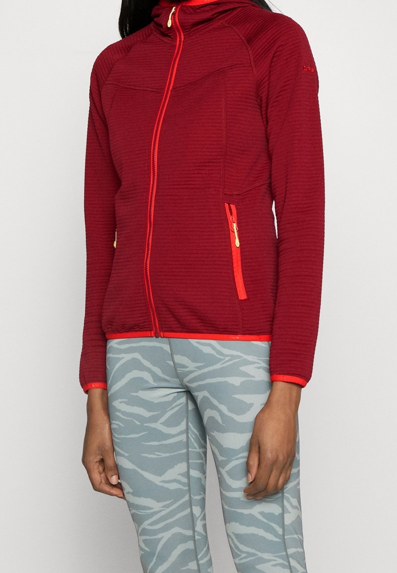 Personne portant une veste rouge zippée texturée avec des tirettes de fermeture éclair orange et des leggings gris clair avec un motif ondulé.