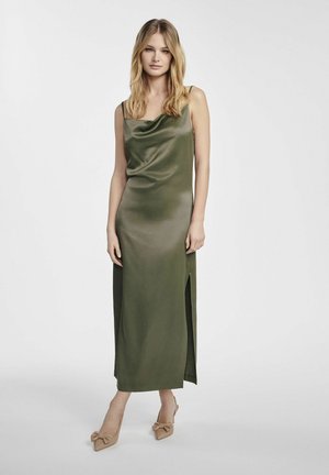 Zielona satynowa sukienka o dekolcie w łezkę i bocznych rozcięciach; posiada ramiączka spaghetti i dopasowaną sylwetkę, zestawiona z beżowymi szpilkami.