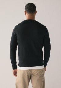 Fekete kötött pulóver hosszú ujjakkal, kerek nyakkivágással és bordázott mandzsettákkal, fehér ing fölött viselve, világos bézs nadrággal párosítva.