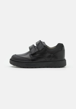 Chaussure décontractée en cuir noir avec deux bandes Velcro, zones texturées au niveau des orteils et du talon, et semelle épaisse en caoutchouc noir.