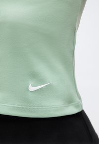 Κοντινό πλάνο ενός ανοιχτοπράσινου αθλητικού πουκαμίσου με λευκό λογότυπο Nike Swoosh κοντά στο τελείωμα, φορεμένο πάνω από μαύρο παντελόνι προπόνησης.