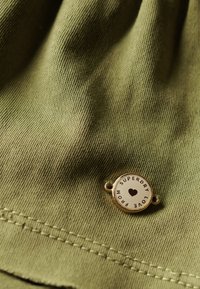 Olivgrüner Stoff mit strukturierter Oberfläche, versehen mit einem runden goldenen Anhänger, auf dem „Superdry Love“ und ein Herzsymbol zu sehen sind.