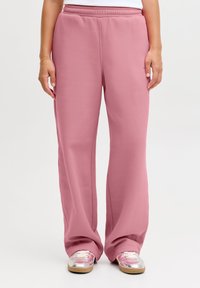 Růžové tepláky s volným střihem, elastickým pasem a hladkou strukturou. Nošeny s metalickými stříbrnými teniskami s růžovým vzorem.