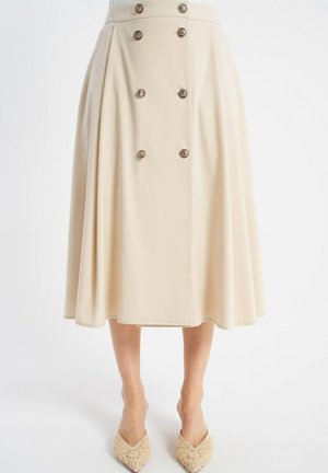 Beżowa spódnica midi z podwójnym rzędem guzików z przodu, plisowane boki, noszona z beżowymi, tkanymi balerinami z ostro zakończonym noskiem na białym tle.