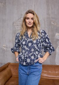 Blouse bleu marine à motif floral blanc, manches bouffantes et col en V. Associée à un jean bleu taille haute et une bague en argent. Fond texturé.
