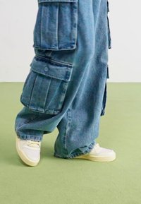 Wide blue denim cargo pants with multiple pockets, paired with white sneakers. Texture is smooth; overall design is relaxed-fit.