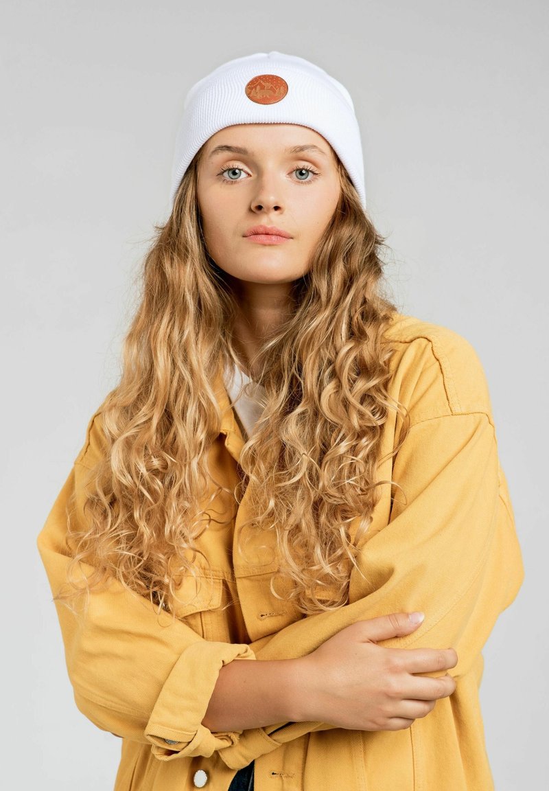 Biała dziana czapka z okrągłą brązową skórzaną naszywką, noszona przez osobę w żółtej, oversize'owej kurtce, ukazująca teksturę i kontrast kolorów.