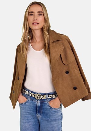 Femme aux longs cheveux blonds portant une chemise blanche, un jean bleu, une ceinture imprimée léopard et une veste en daim marron drapée sur les épaules.