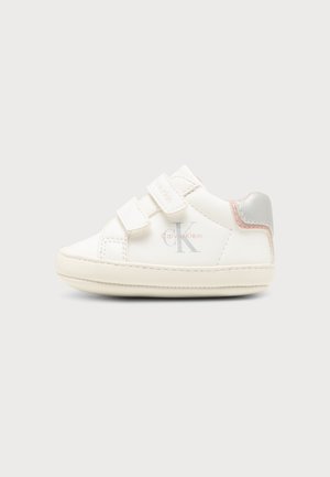 Zapatilla blanca para niño pequeño con doble cierre de velcro, logo de Calvin Klein en el lateral y detalle plateado y rosa en el talón sobre fondo liso.