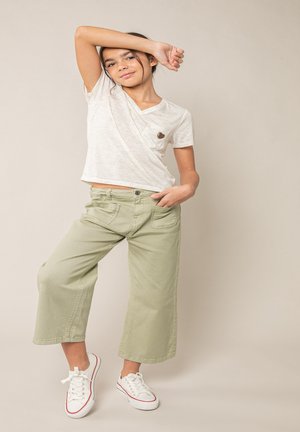 Fille portant une chemise blanche à manches courtes avec un écusson en forme de cœur, un pantalon large vert clair et des baskets blanches, posant avec un bras levé.