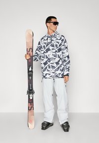 Snowboarder che indossa un hoodie a motivi bianchi e neri, pantaloni grigi chiari e stivali neri, tiene un sci con attacchi, su uno sfondo semplice.