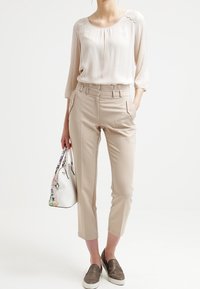 Lys beige bluse med blonde detaljer, tapered beige bukser og grå slip-on sko. Hvid håndtaske med blomsterdetaljer fuldender outfittet.
