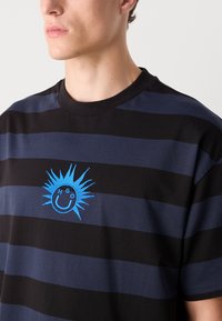 Black and navy striped t-shirt with a ribbed collar. Features a blue embroidered sun graphic with a smiley face and the text "H G O."