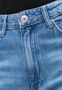 Κοντινό πλάνο της μέσης ενός τζιν παντελονιού γαλάζιου χρώματος με μεταλλικό κουμπί και θηλιές για ζώνη, πάνω σε μαύρο υφασμάτινο φόντο.