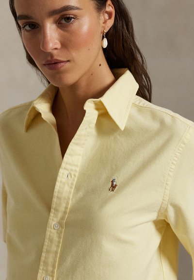 Frau mit langen braunen Haaren trägt kleine weiße Tropfenohrringe und ein blassgelbes Hemd mit Knöpfen und einem kleinen bestickten Polo-Spieler-Logo.