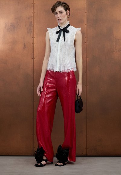 Haut en dentelle blanche avec bords volantés et nœud noir au niveau de l'encolure, associé à un pantalon large rouge scintillant à sequins et des sandales ornées de fleurs noires.