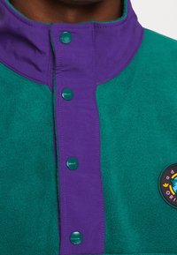Fleecetröja med grön kropp, lila krage och kontrasterande knappar. Inkluderar en rund logotypmärke med färgglada designdetaljer.