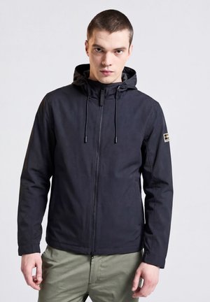 Mann trägt schwarze Kapuzenjacke mit Reißverschluss und olivgrüne Hose, steht vor hellem einfarbigem Hintergrund.