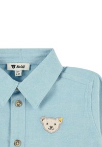 Camicia di jeans azzurra con colletto morbido, caratterizzata da un patch ricamato a forma di faccia d'orso sul petto e bottoni marroni.