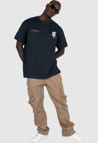Black cotton t-shirt with white graphic detail on left chest, paired with beige cargo pants featuring multiple pockets and cream sneakers.