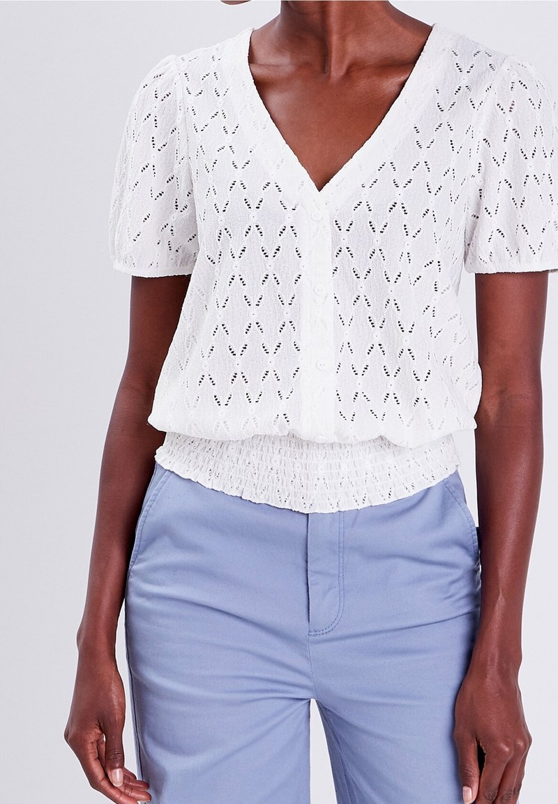 Femme portant un chemisier blanc texturé à manches courtes avec des boutons et un pantalon taille haute bleu clair, debout sur un fond uni.