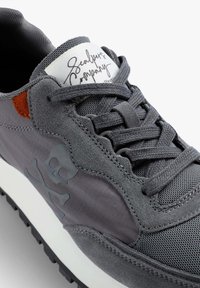 Graue Sneaker mit einer Kombination aus verschiedenen Materialien, darunter Wildleder und Mesh. Er hat ein Schullogo, strukturierte Akzente und eine weiße Zwischensohle.