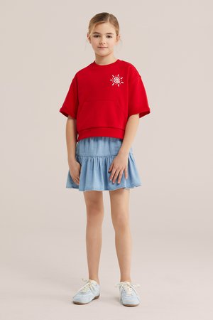 Noor tüdruk, kes kannab punast lühikese varrukaga topi päikese graafikuga, helesinist volditud seelikut ja valgeid tossusid, seisab neutraalse tausta ees.