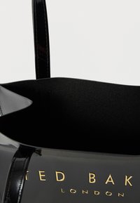 Fekete lakkbőr bevásárlótáska sima textúrával, aranyszínű "TED BAKER LONDON" logóval és két vékony, fényes fogóval.