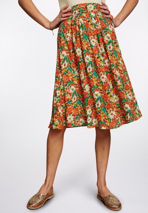 Jupe à hauteur du genou avec un motif floral aux couleurs vives orange, blanc et vert, portée avec des chaussures plates tissées dorées et un haut beige.