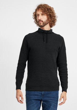 Schwarzer Strickpullover mit hohem Kragen, Kordelzugakzenten und gerippten Bündchen. Strukturiertes Material und taillierte Schnittform. Getragen über blauen Jeans.