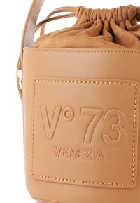 Sac seau en cuir brun clair avec une fermeture à cordon, logo embossé et coutures détaillées. Doublure en daim doux visible sur le dessus.